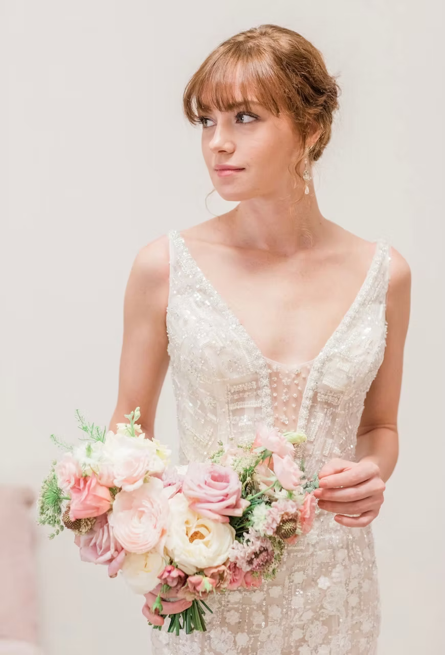 Bride in a beaded wedding dress, holding a pink and lavender bouquet, looking to the side.