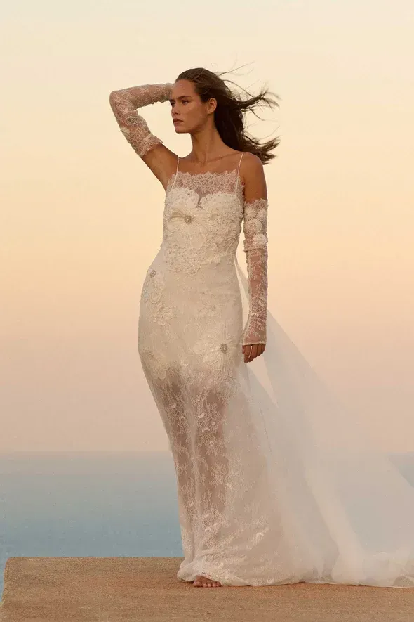 Woman in wedding dress with sheer sleeves, overlooking ocean at sunset.