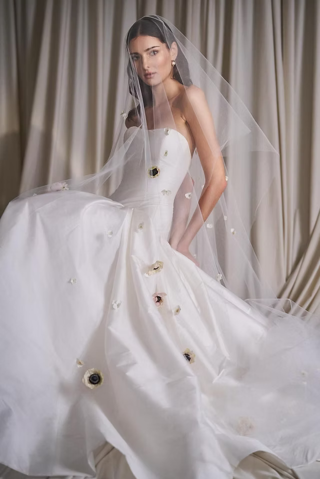 Bride in white strapless gown, veil with floral accents, seated, cream backdrop.
