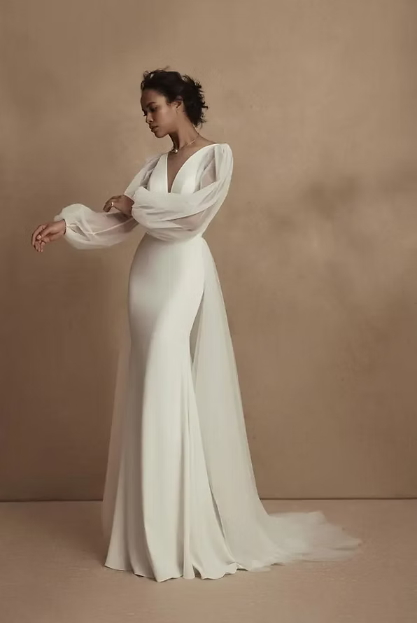 Woman in a white wedding dress with long sheer sleeves and a train, posing in a studio.