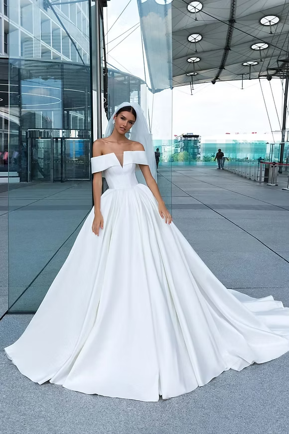 Bride in a white, off-shoulder gown, with a long train, poses outdoors near modern glass building.