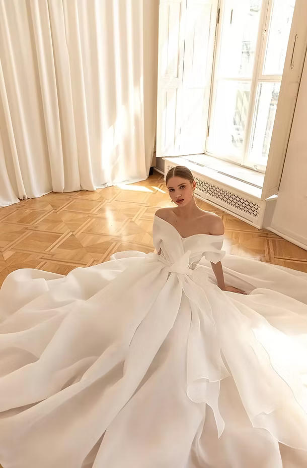 Woman in a white off-the-shoulder wedding gown sits on wooden floor near window.