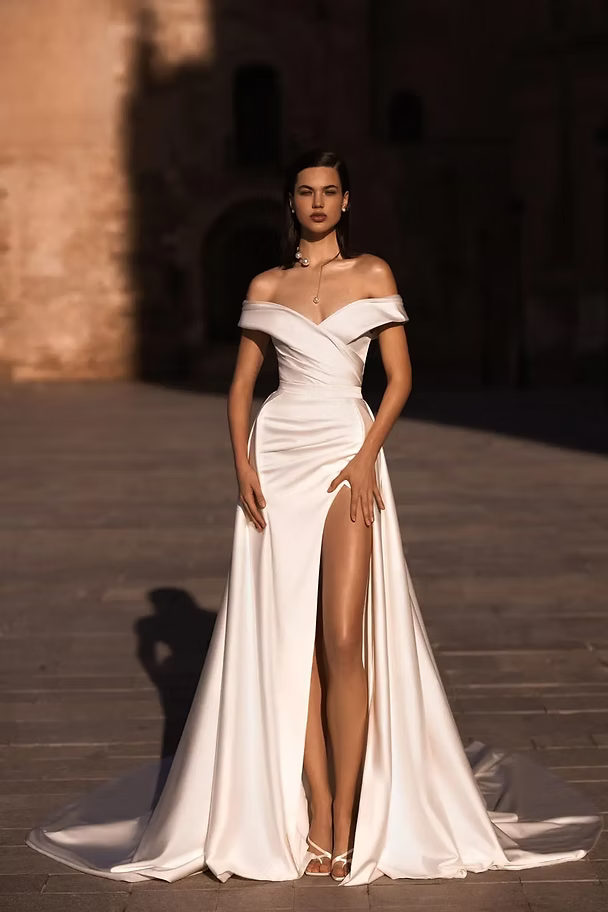 Woman in white off-the-shoulder gown with high slit, posing outdoors in sunlight.