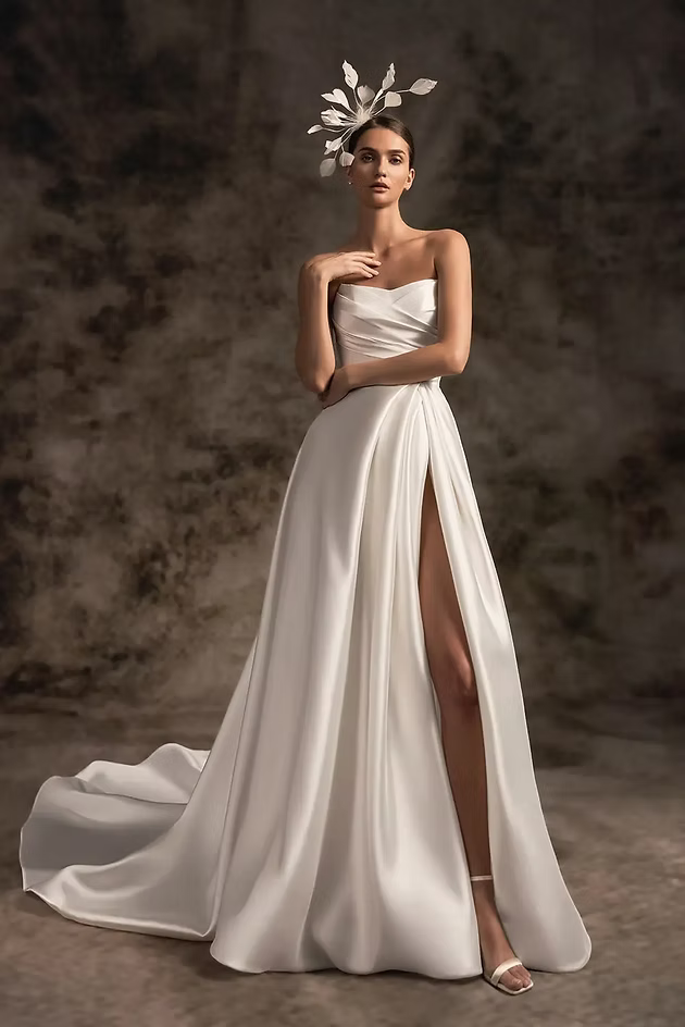 Woman in white strapless wedding dress with high slit, wearing a headpiece; posing.