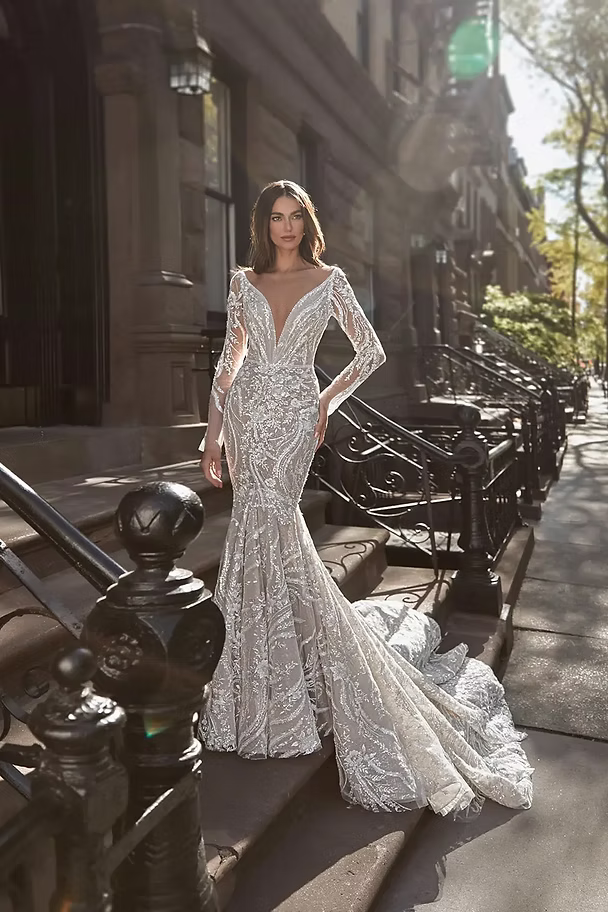 Woman in a shimmering white mermaid gown, poses on steps with a long train, outdoor setting.