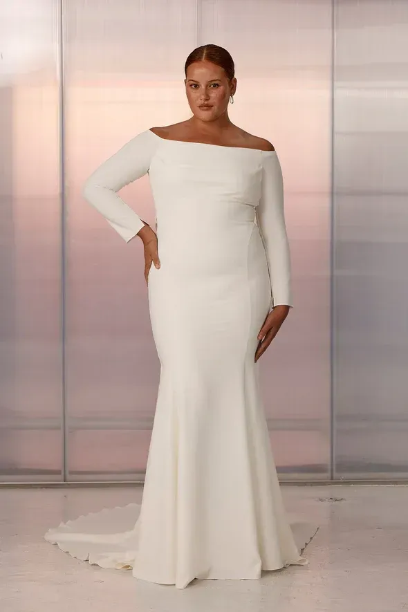 Woman in an off-the-shoulder white wedding dress with a mermaid skirt, posing in front of a reflective wall.