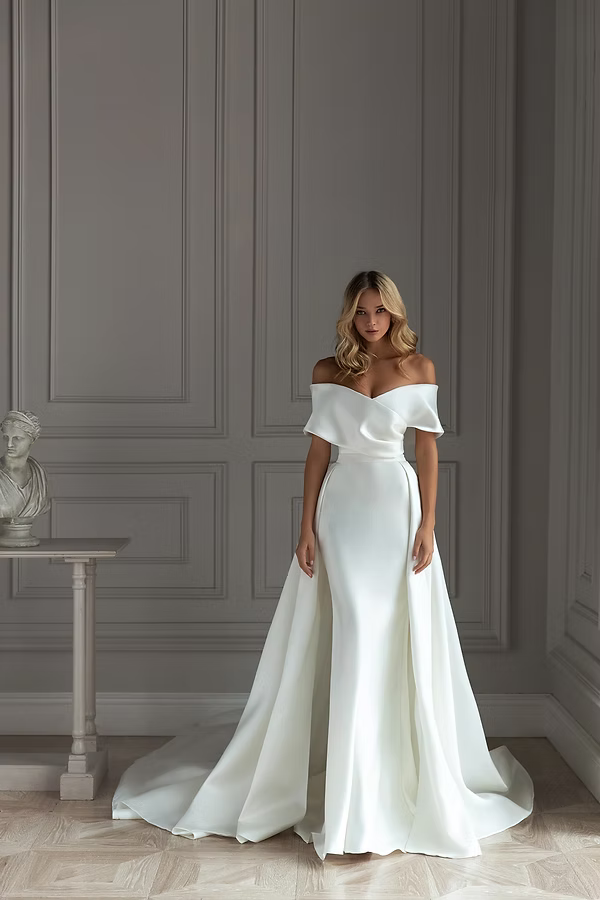 Woman in white off-the-shoulder wedding dress with long train, standing indoors with a gray wall.