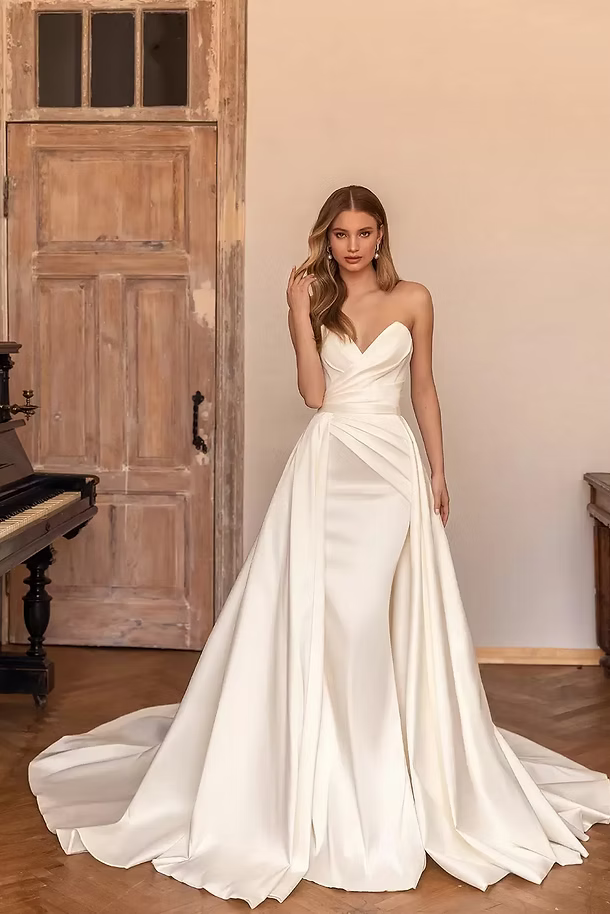 Woman in a white strapless wedding dress with a long train, standing in a room with a piano.