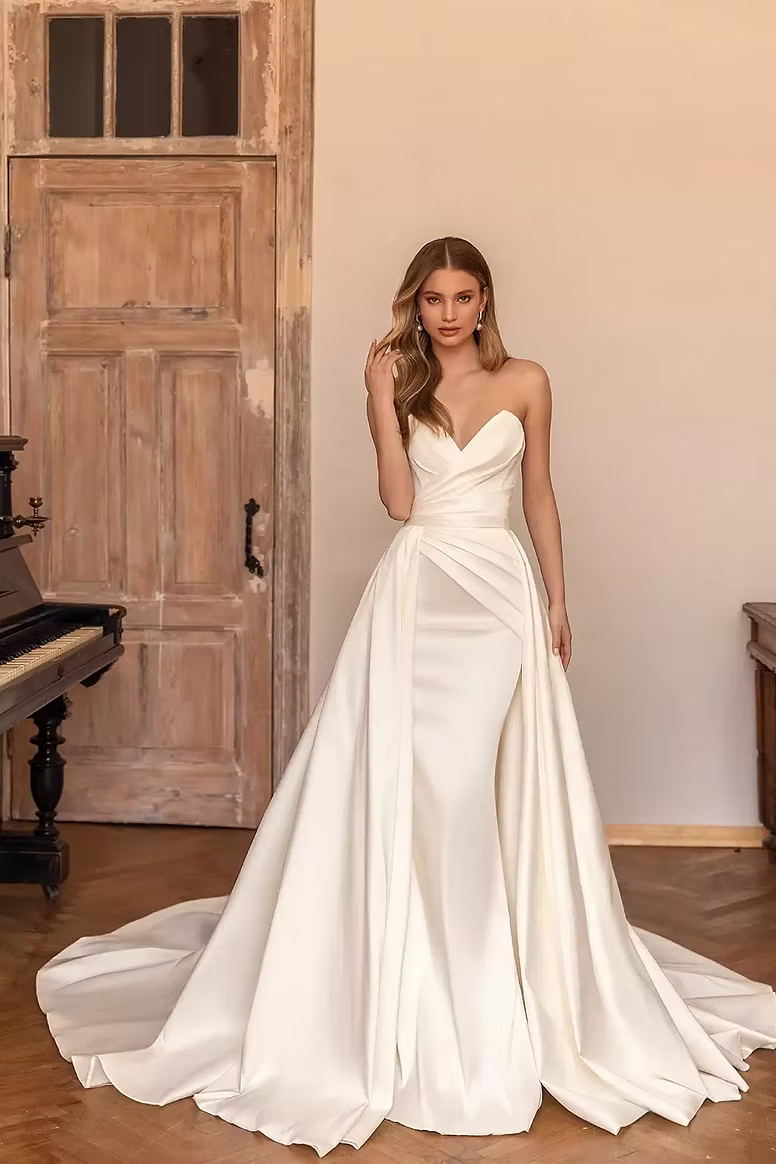 Woman in a white strapless wedding dress with a long train, standing indoors.