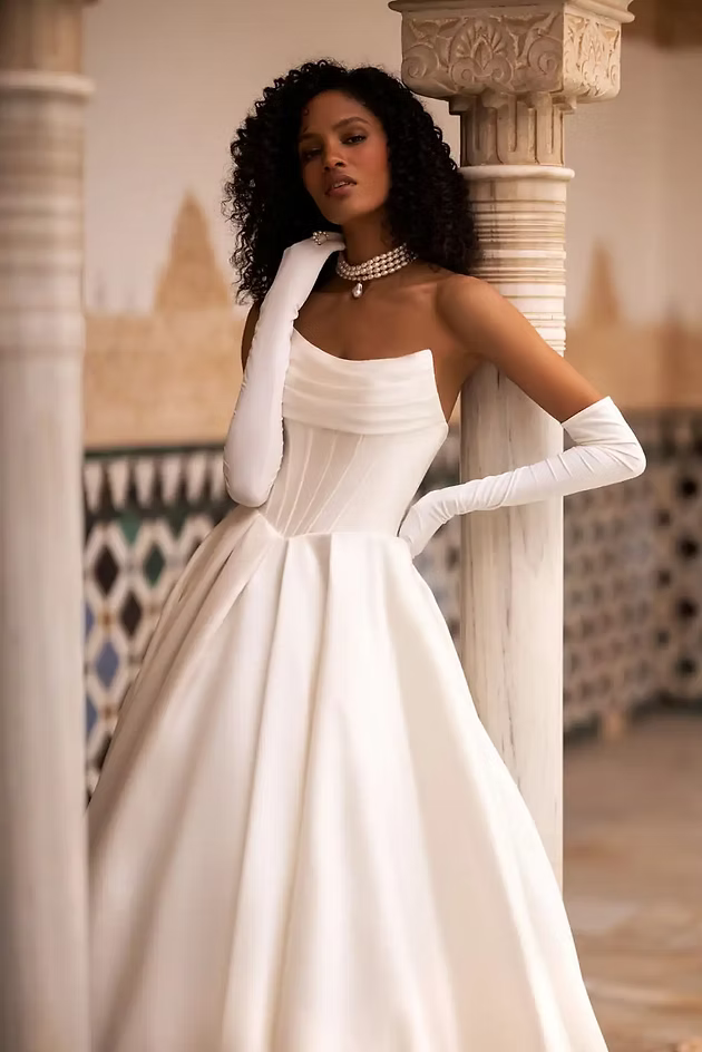 Woman in white gown and gloves, leaning against column in ornate building.