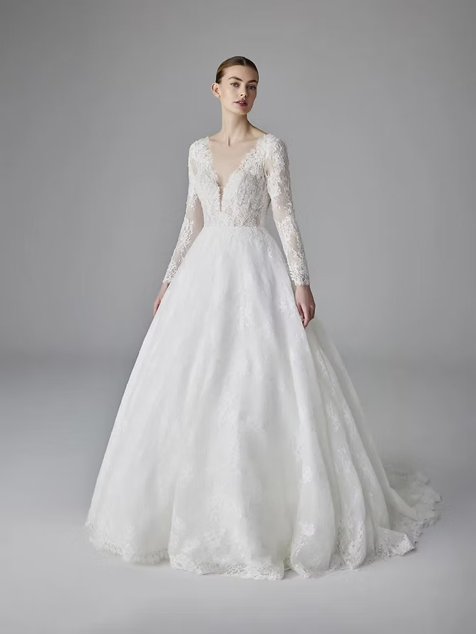 Woman in a white wedding dress with a plunging neckline and long sleeves, standing in a studio.