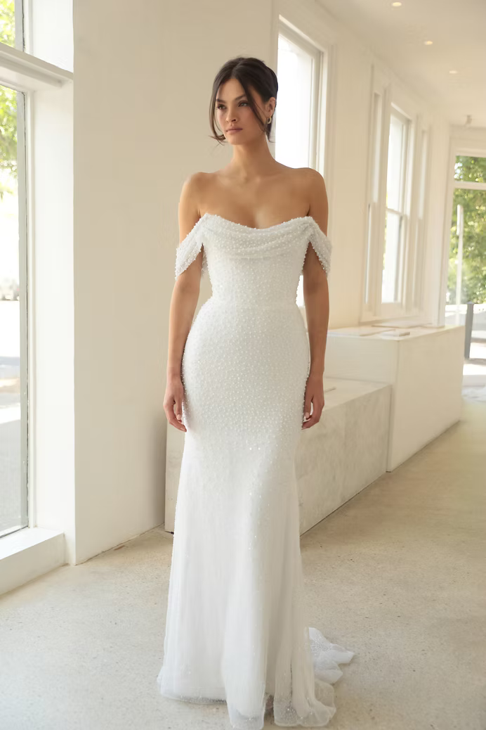 Woman in white off-the-shoulder wedding dress, standing in well-lit room.
