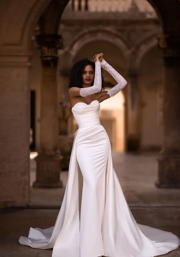 Woman in a white strapless wedding gown with detachable train and lace gloves, posing outdoors.