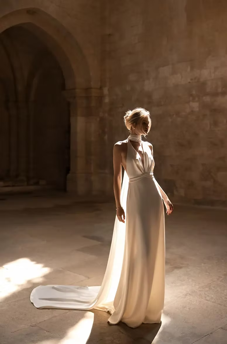 Woman in white gown stands in sunlit, arched hall; train of dress trails behind her.
