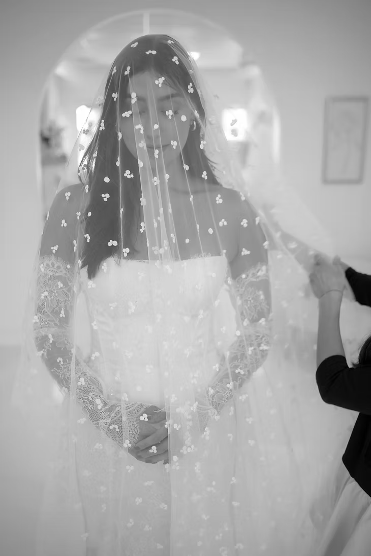 Bride in white dress with veil being adjusted, standing in bright room.