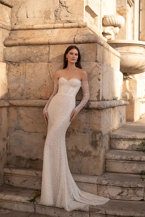 Woman in a sparkling white, strapless wedding dress with sheer sleeves, leaning against a stone building.
