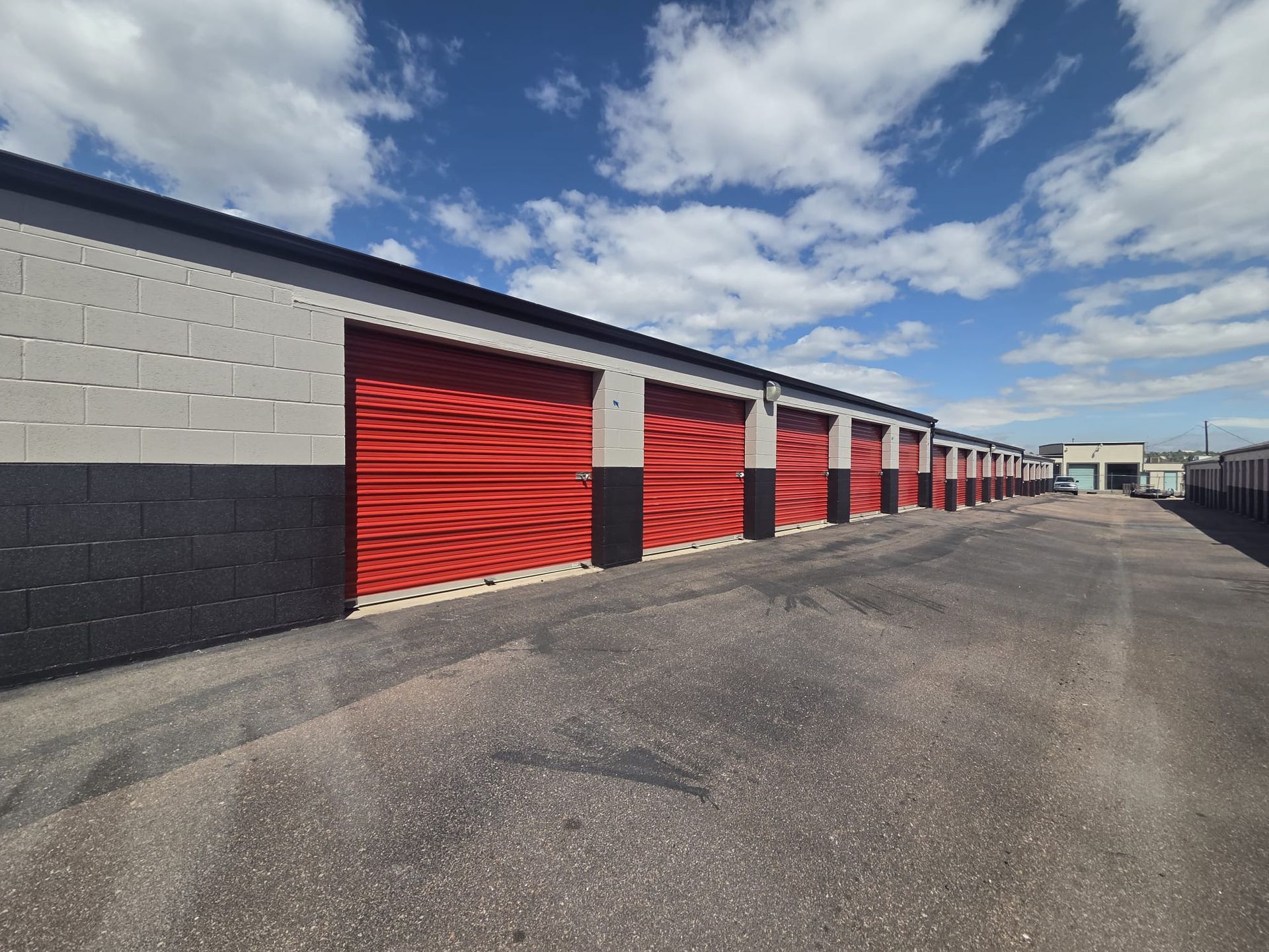 Row of red storage unit doors along a paved lane under a blue sky with clouds