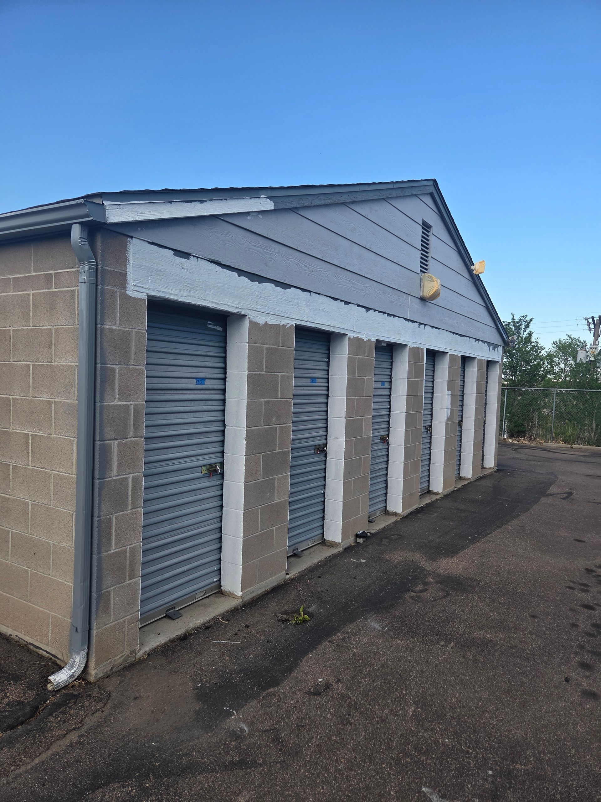 Row of gray storage unit doors on a cinderblock building beside a gravel lot under a blue sky