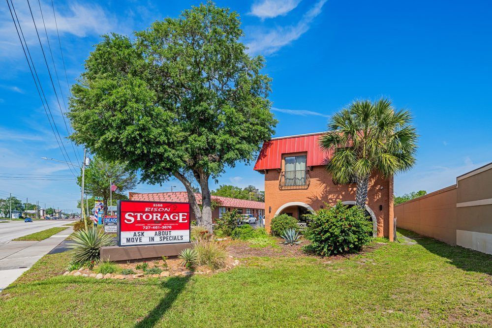 Storage facility sign beside a brick building with red roof, trees, and a grassy roadside lot.