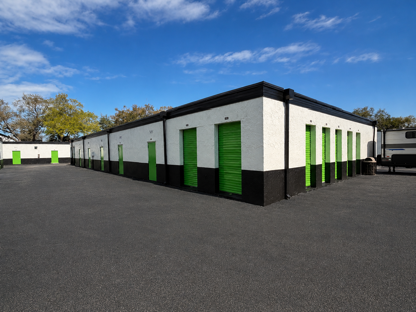 Single-story storage units with green doors on a paved lot under a blue sky.