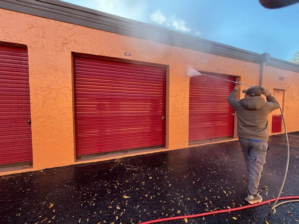 Person pressure-washing red roll-up doors on an orange building exterior.