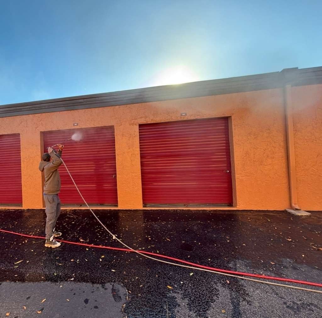 Person pressure-washes a red-orange building with closed red shutters in bright sunlight.