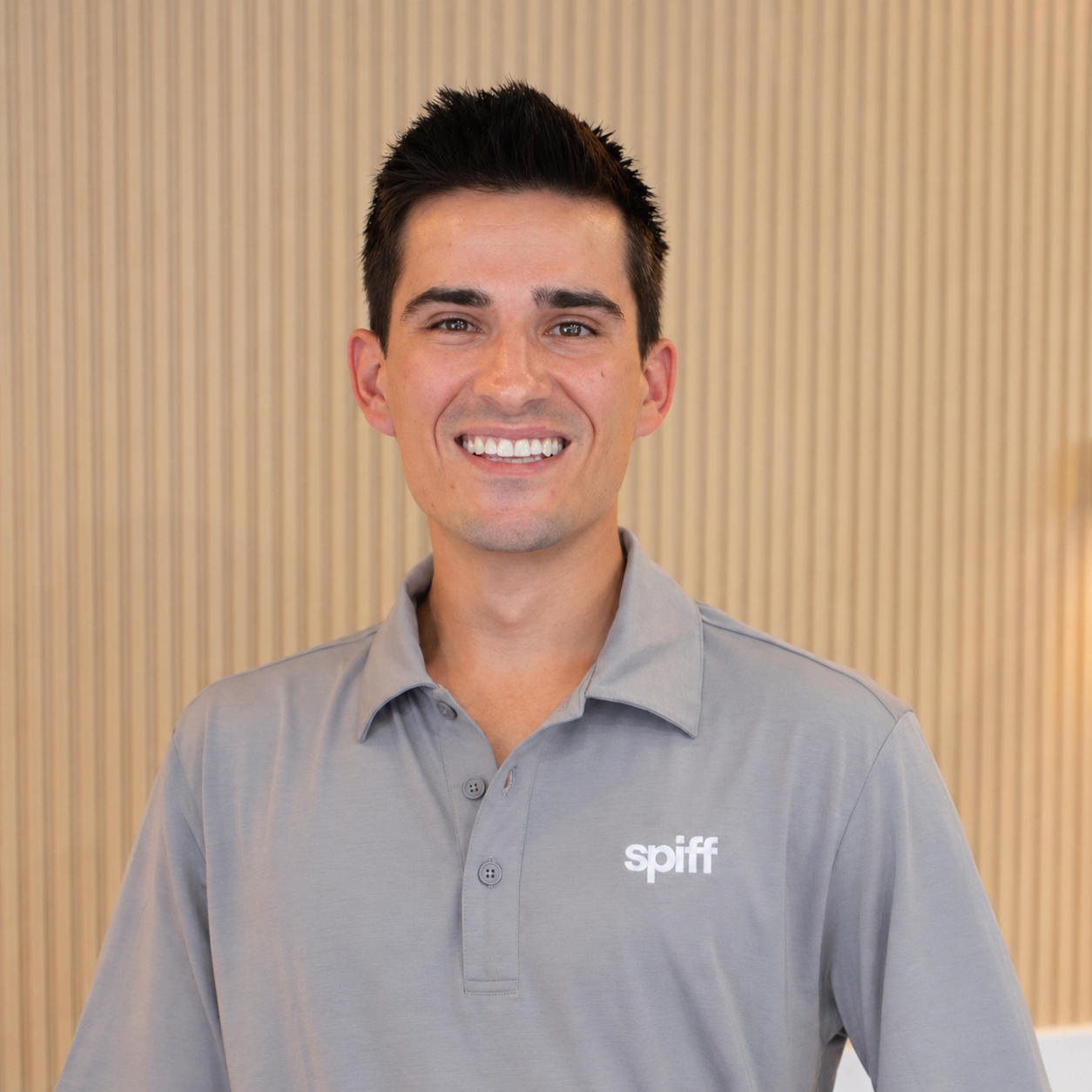 Smiling man in a gray polo shirt standing against a beige striped wall