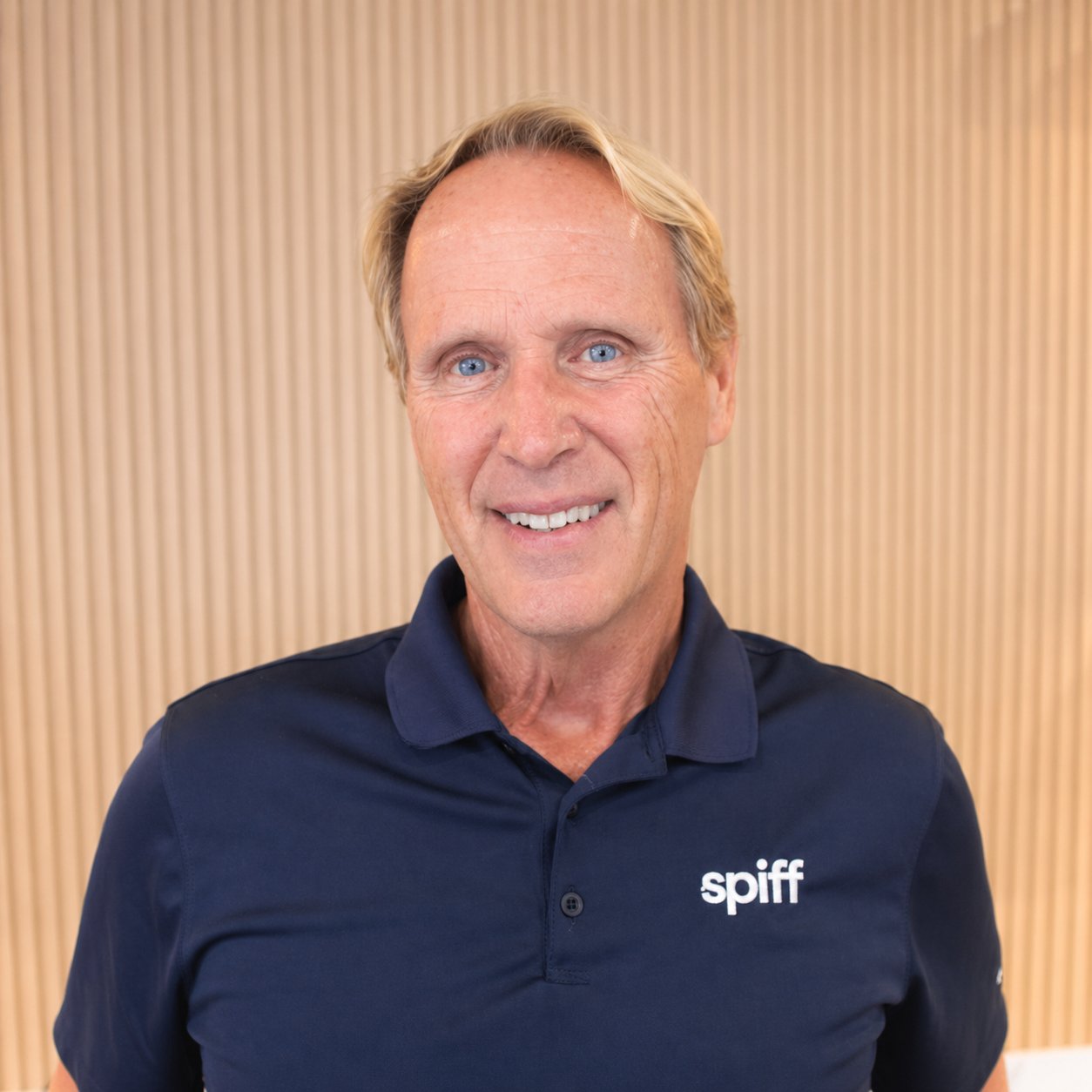 Smiling man in a navy polo shirt with a Spiff logo, standing against a tan striped wall