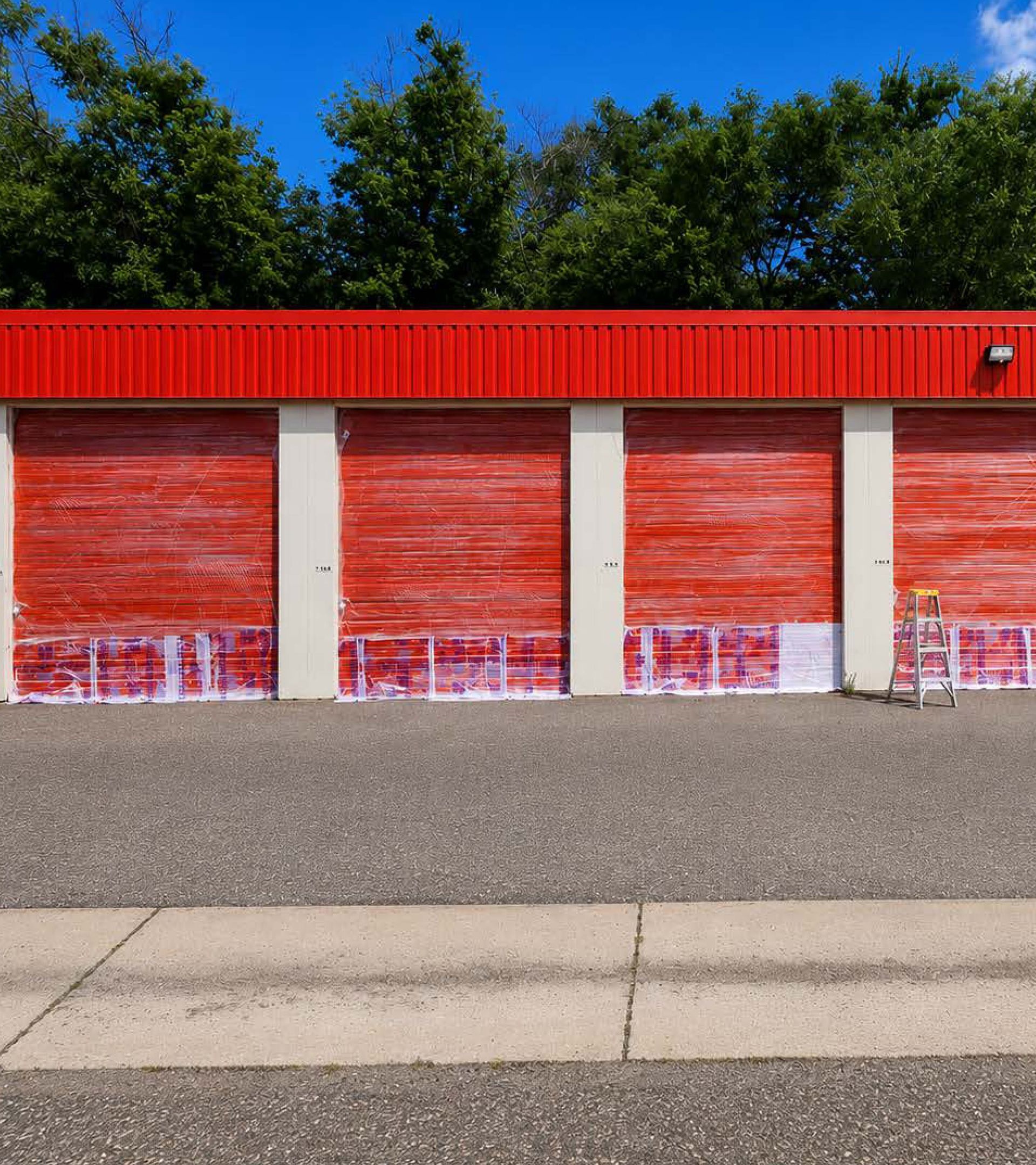 Red storage unit doors in a row with a red roof, set behind a sidewalk and gravel lot under trees.