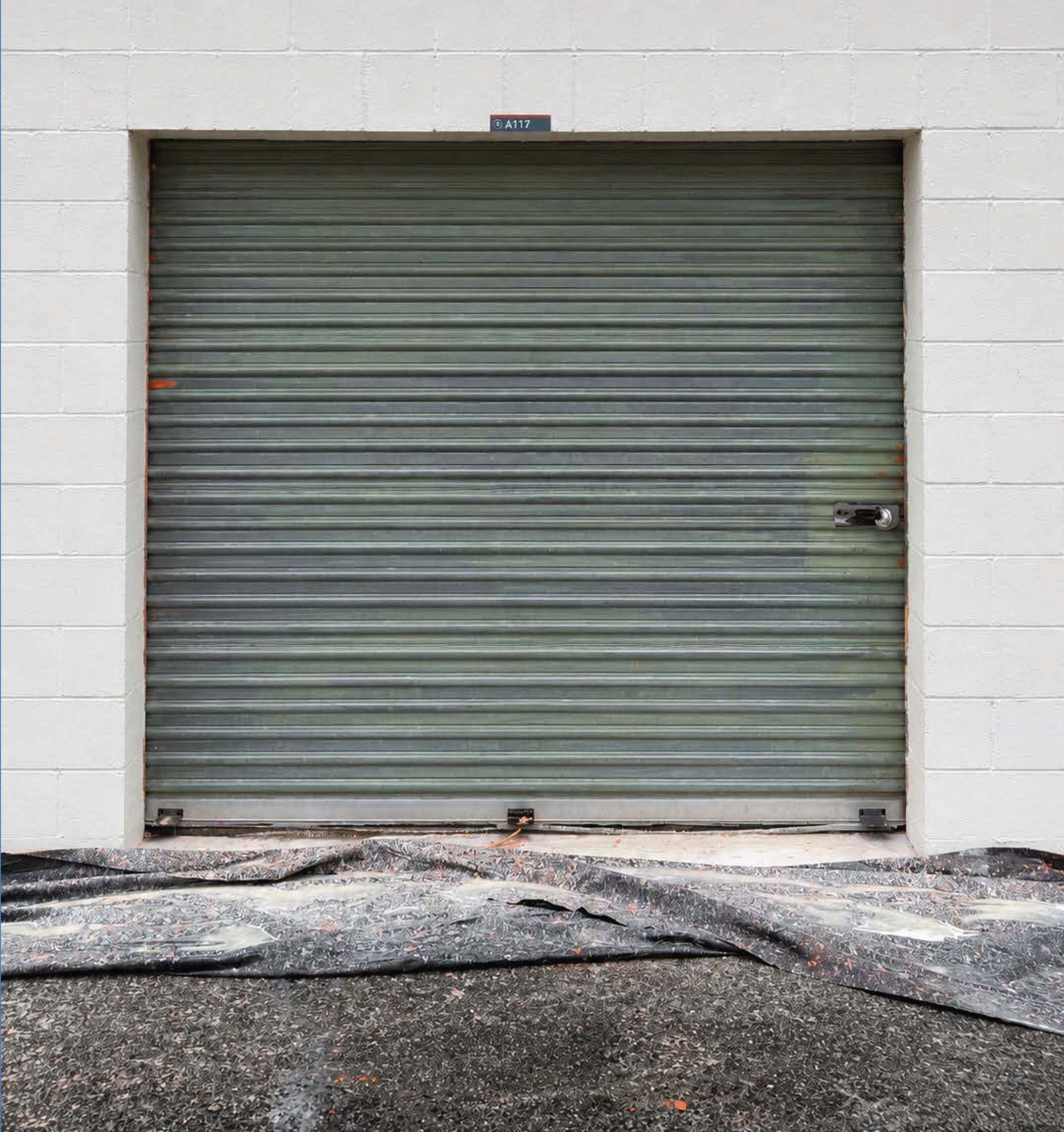 Closed green metal roll-up door set in a white brick wall, with worn pavement in front.