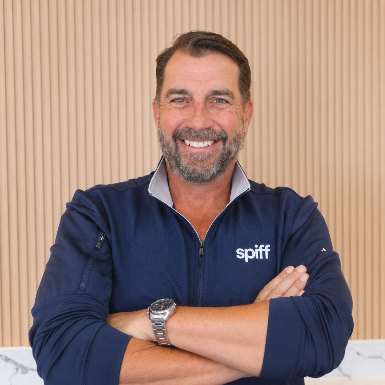 Smiling man in a navy Spiff pullover, arms crossed, standing against a light wood wall