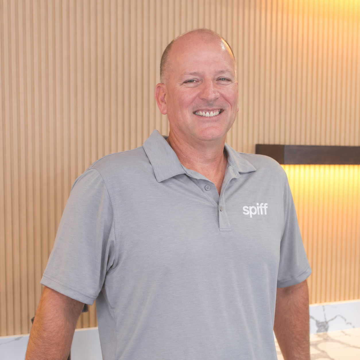 Smiling man in a gray polo shirt stands indoors against a wood-paneled wall.