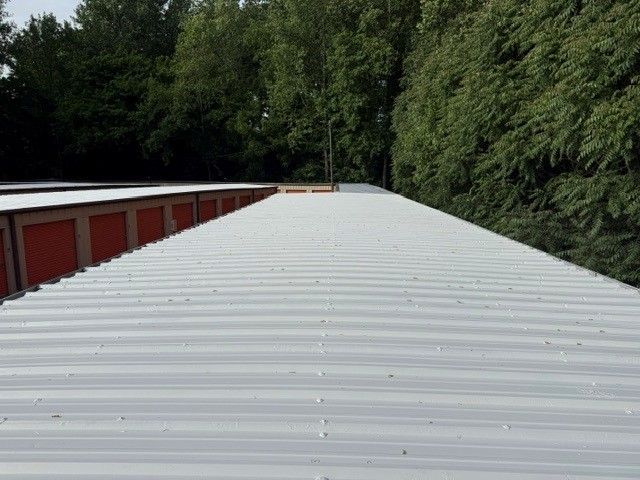 White corrugated roof beside red garages, bordered by dense green trees.