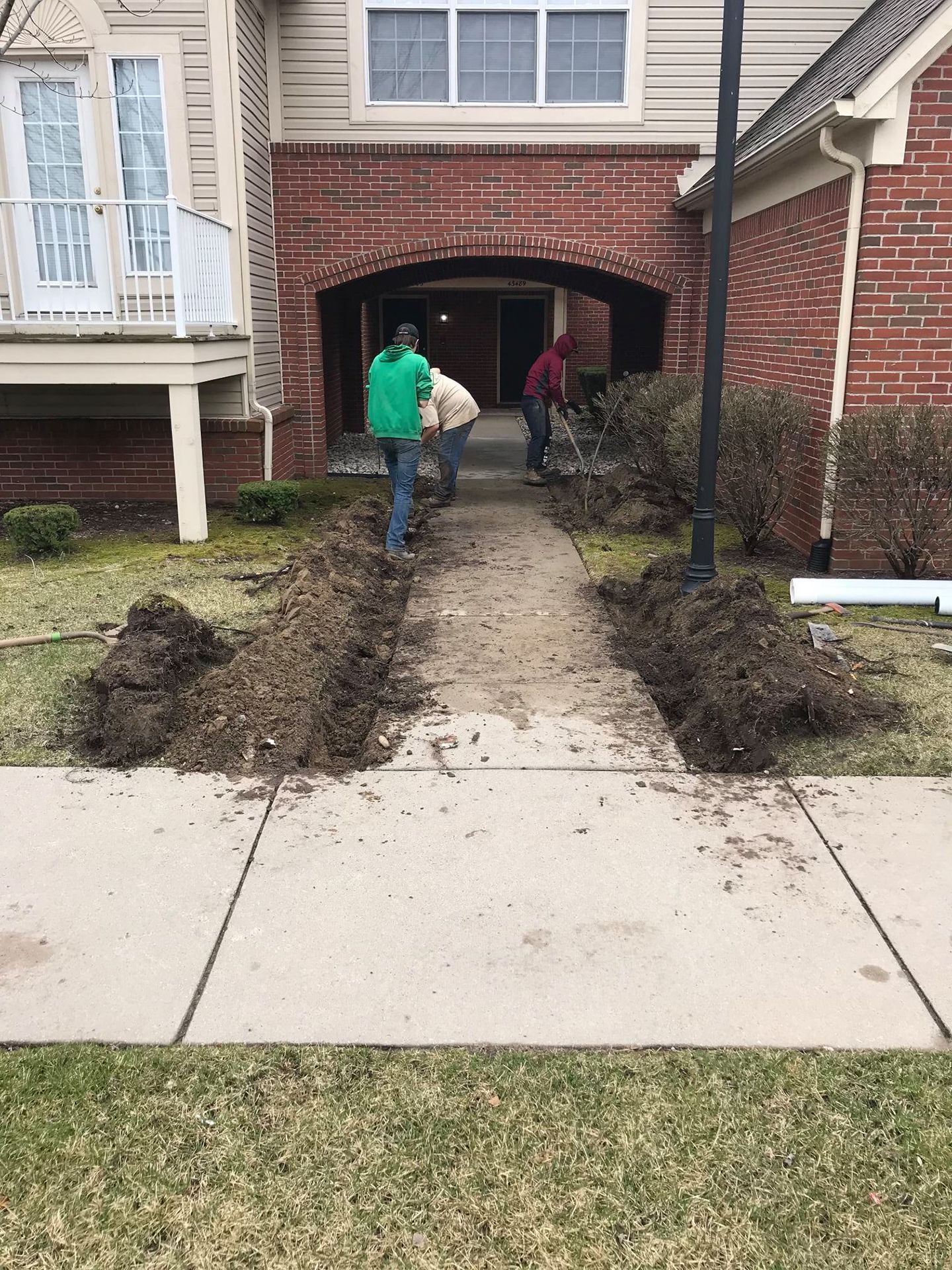 People gardening near an apartment entrance. Dirt piles border a sidewalk.