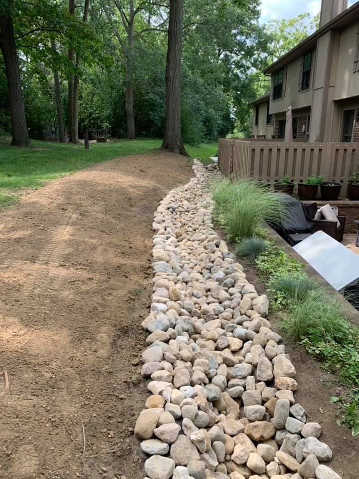 A dry creek bed lined with rocks runs through a grassy yard, near a fence and house.