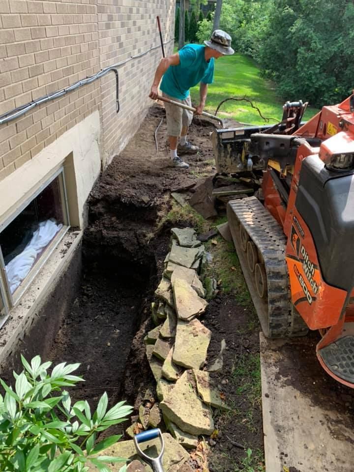 Man using a shovel, digging near a building's foundation with a small excavator.