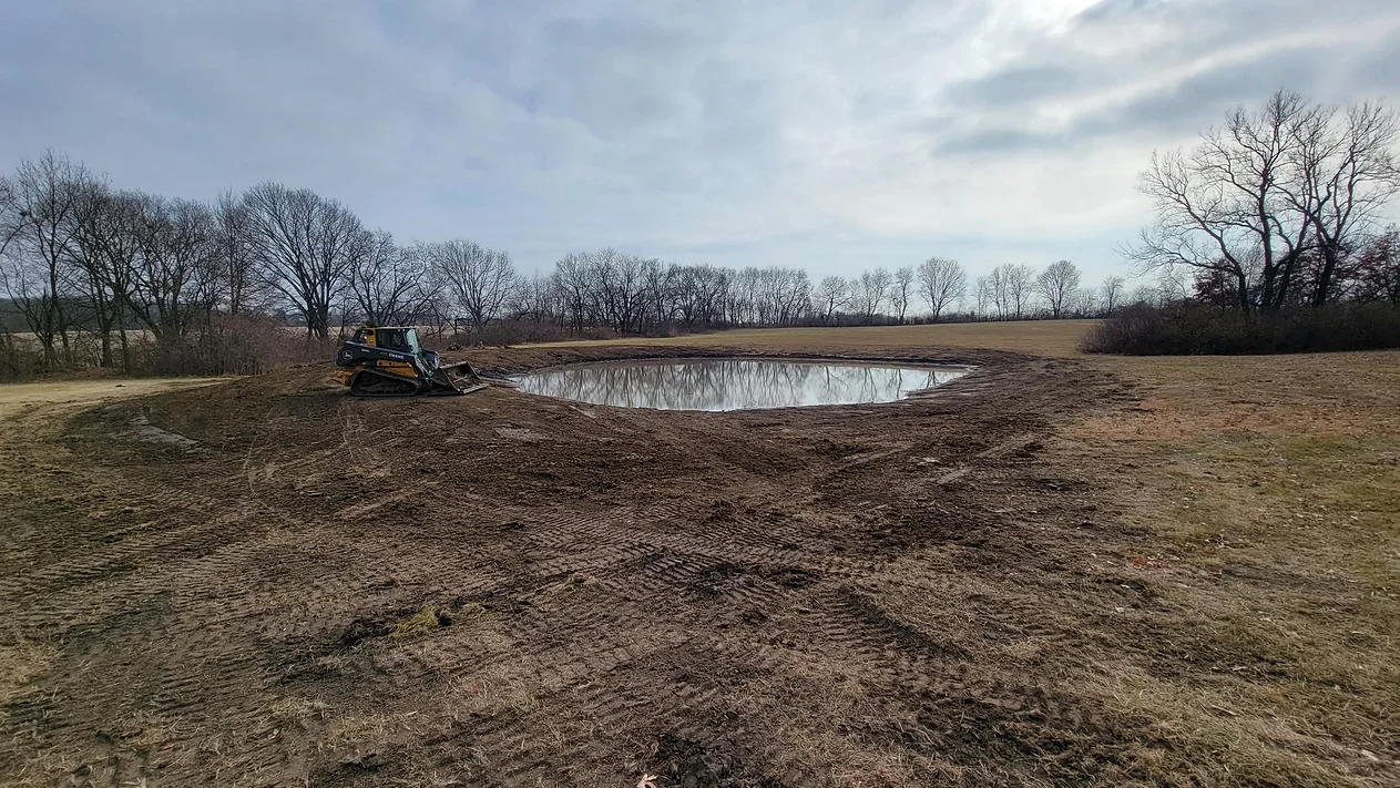 A tractor is working in a field next to a pond.