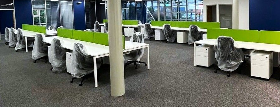 Empty office space with rows of desks, chairs covered in plastic, and green dividers.