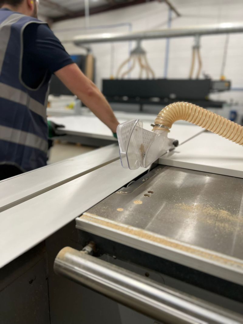 Person in workshop using a saw to cut a white panel, sawdust present.