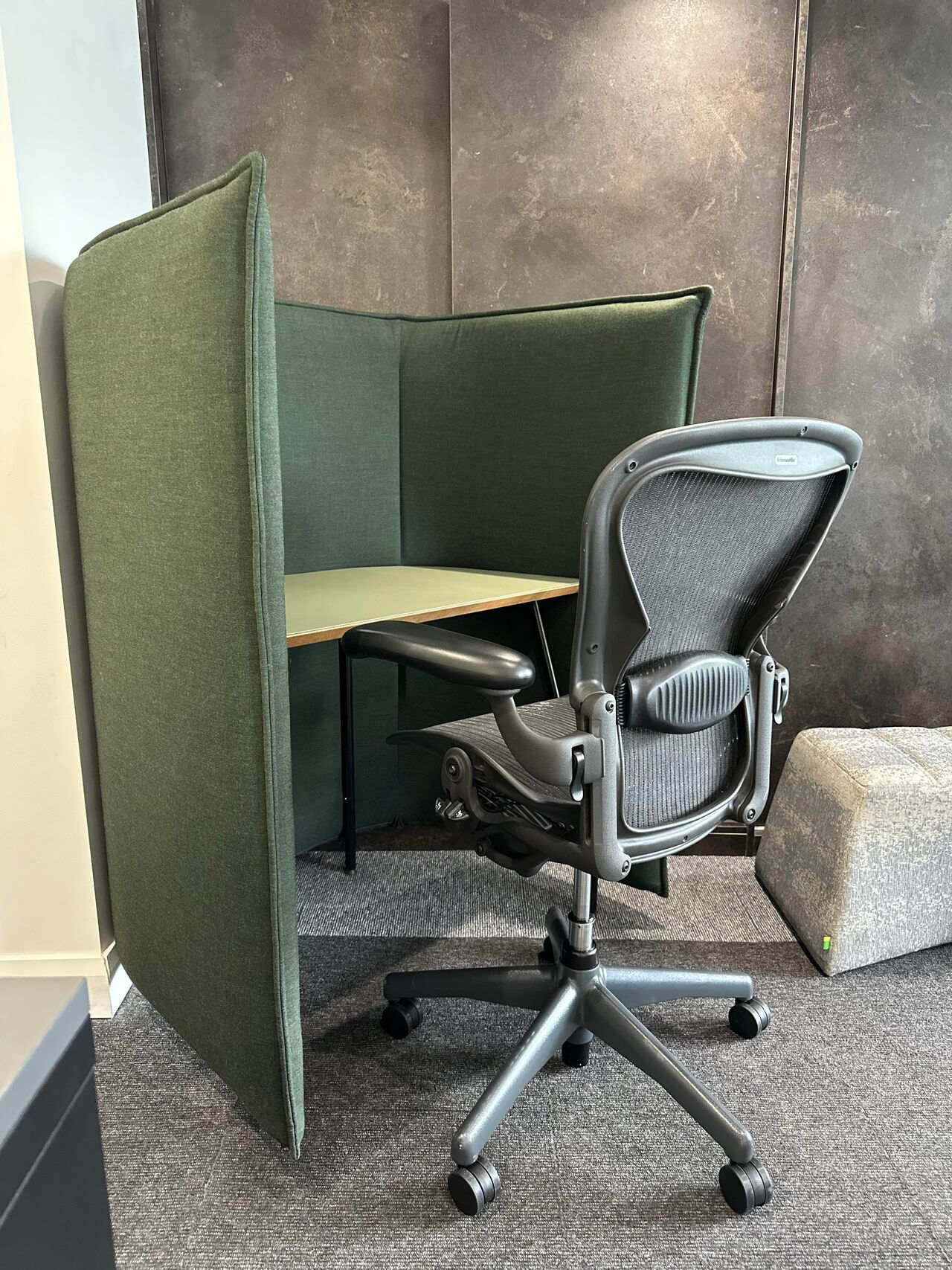 Green-walled workspace with a black office chair and small desk. Light gray carpet, a gray textured wall, and a gray ottoman are visible.