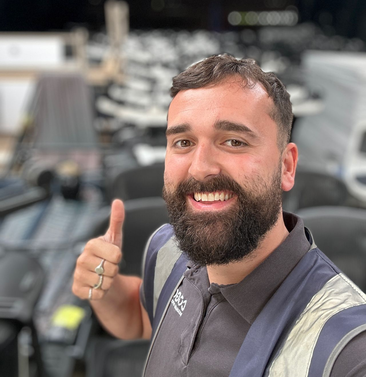 Man with a beard giving a thumbs up, smiling in a store, wearing a vest and polo shirt.