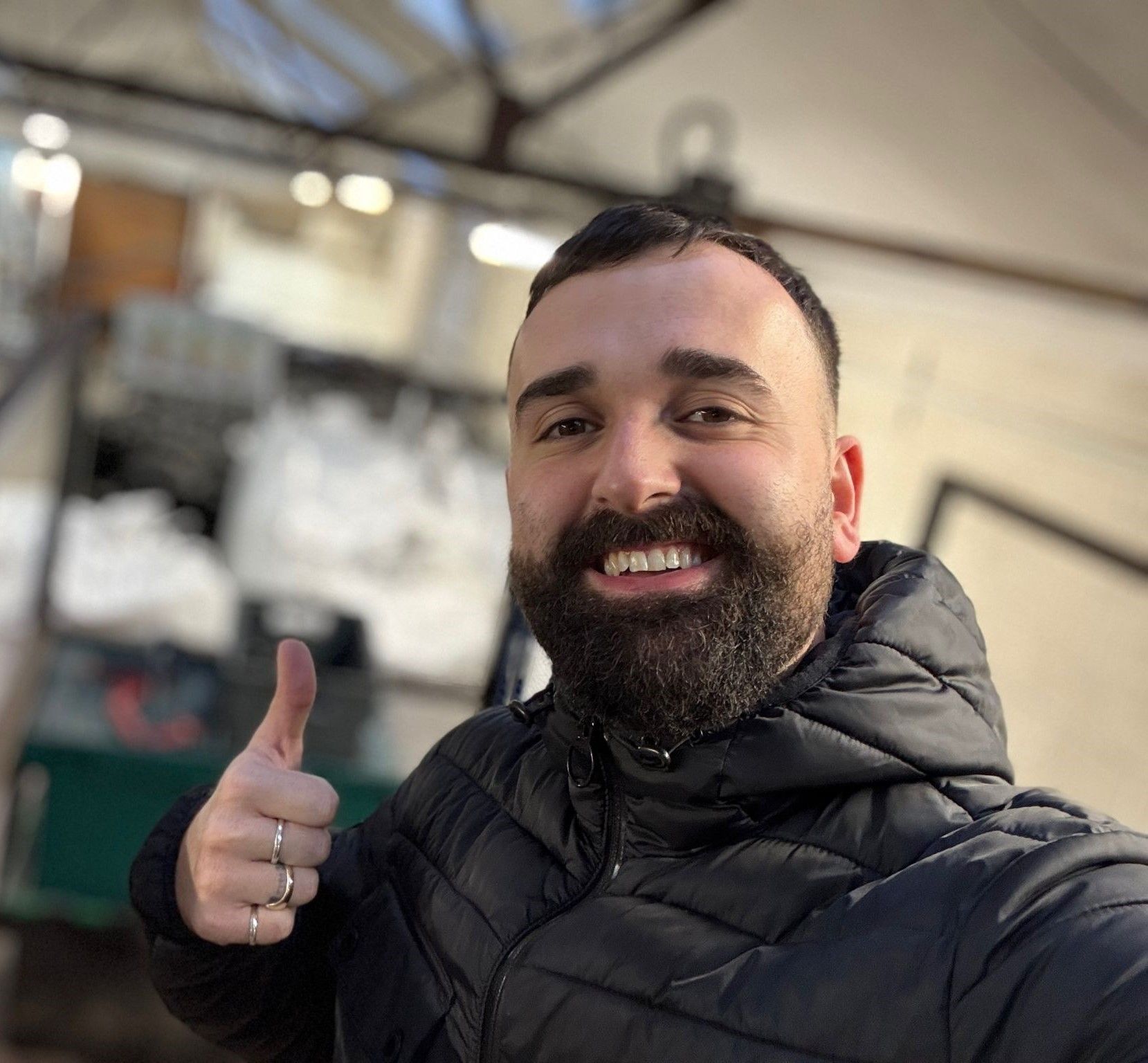 Man with a beard in a black puffer jacket giving a thumbs-up inside a warehouse.