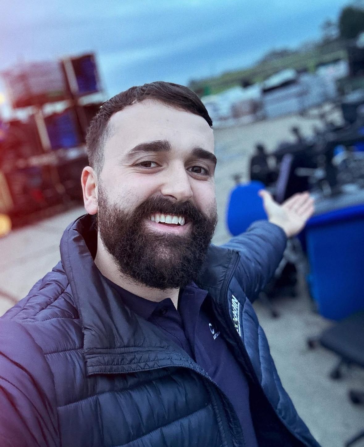 Man with a beard smiles, points towards office chairs in an outdoor setting, wearing a navy jacket.