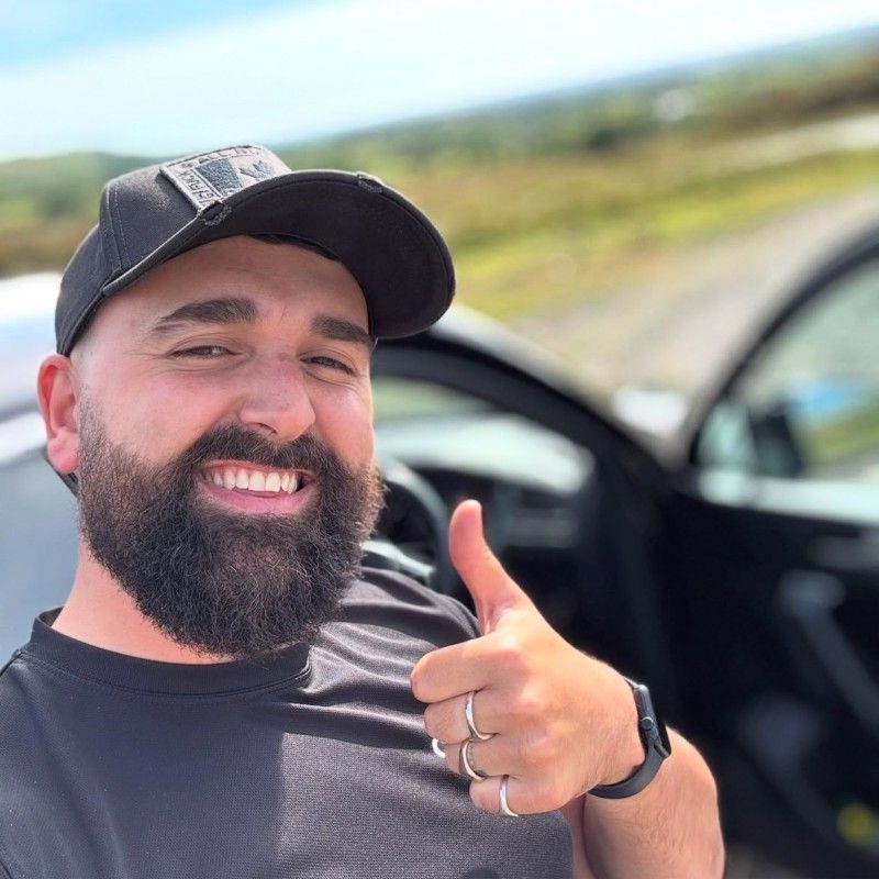 Man with beard, thumbs up, wearing a black cap, smiling, near a car, outdoors.