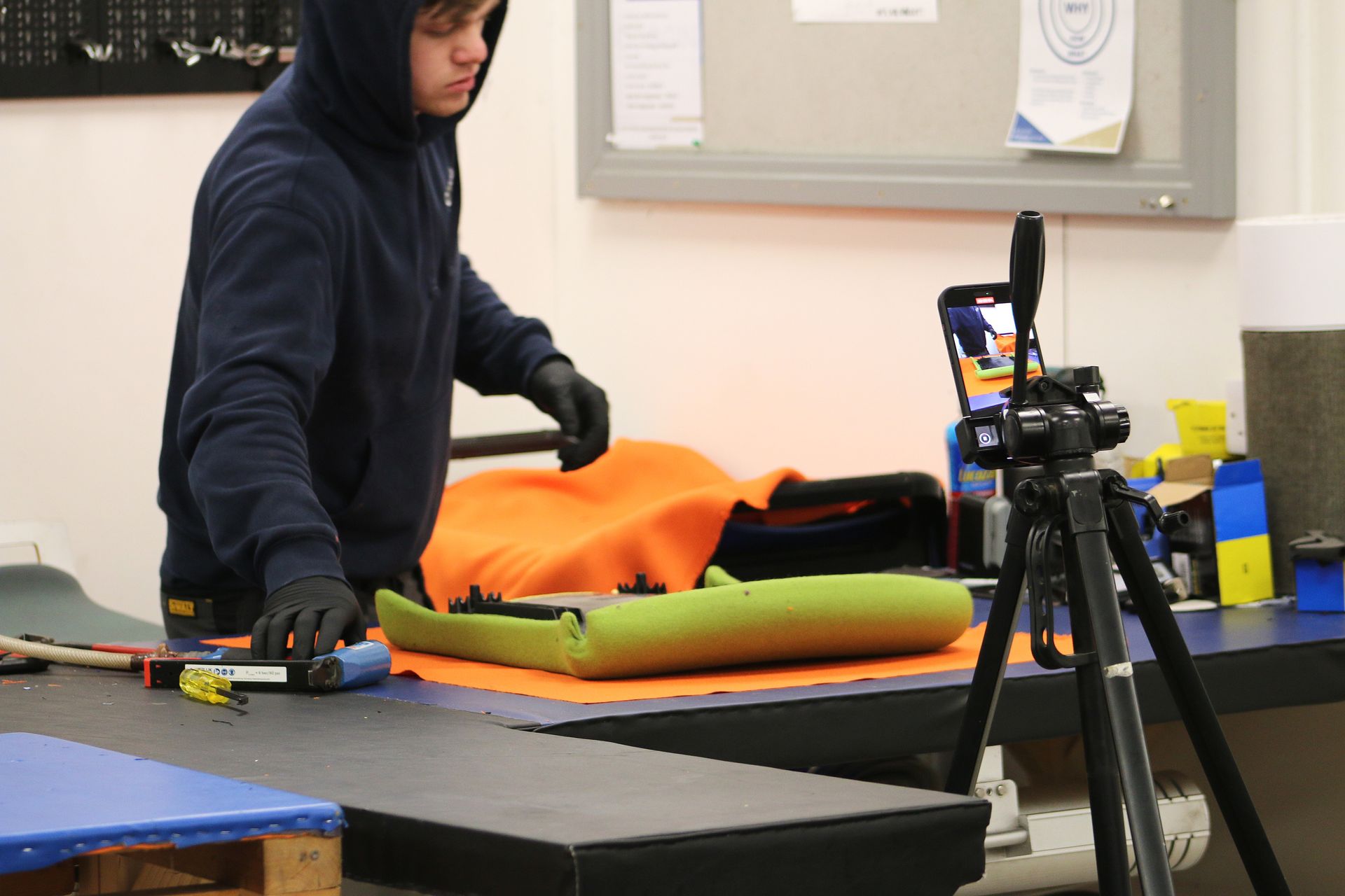 Person in a hoodie, filming car seat repair, using gloves. Brightly lit workshop.