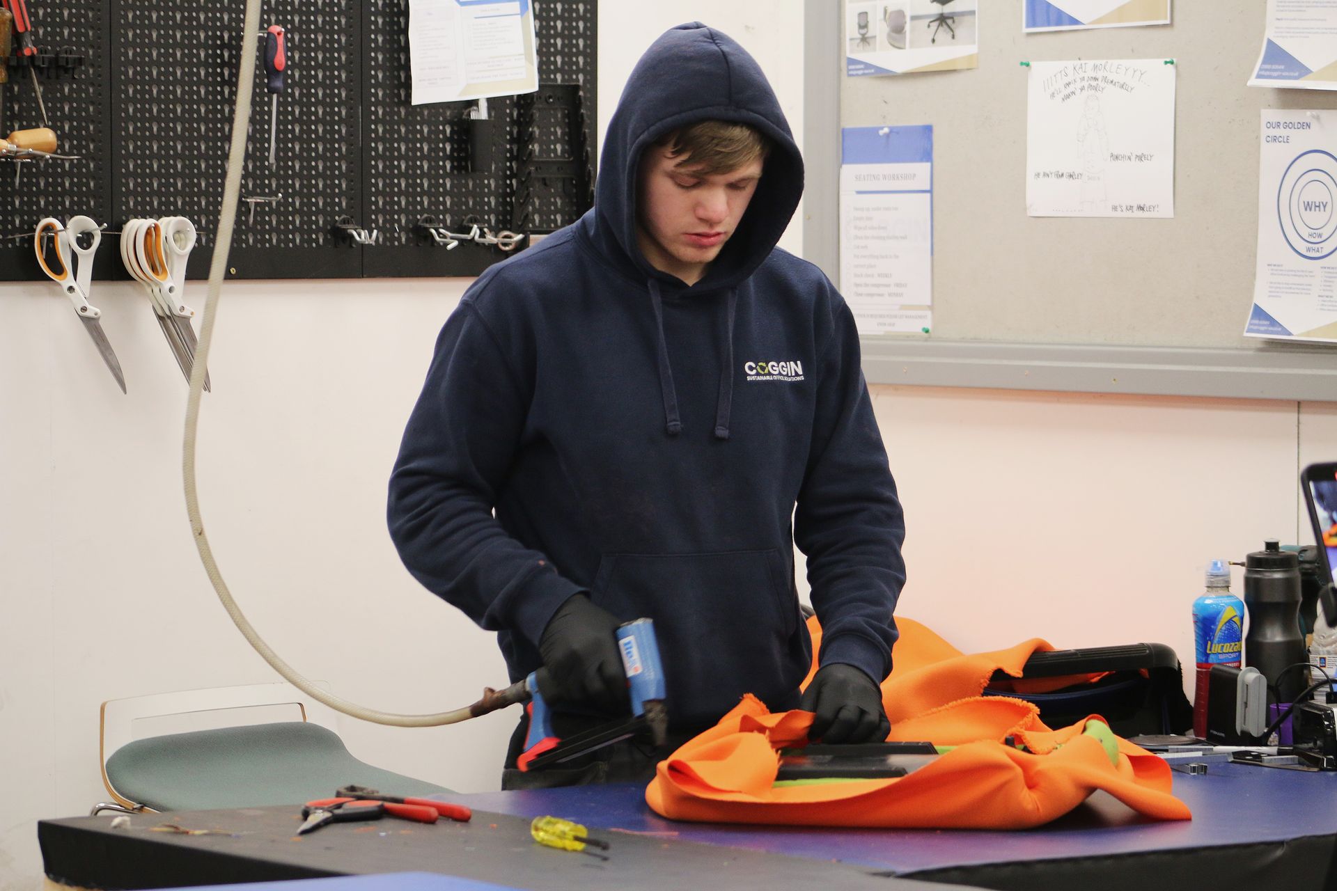 Person in a blue hoodie works on a project at a workbench. Black gloves and orange cloth are visible.