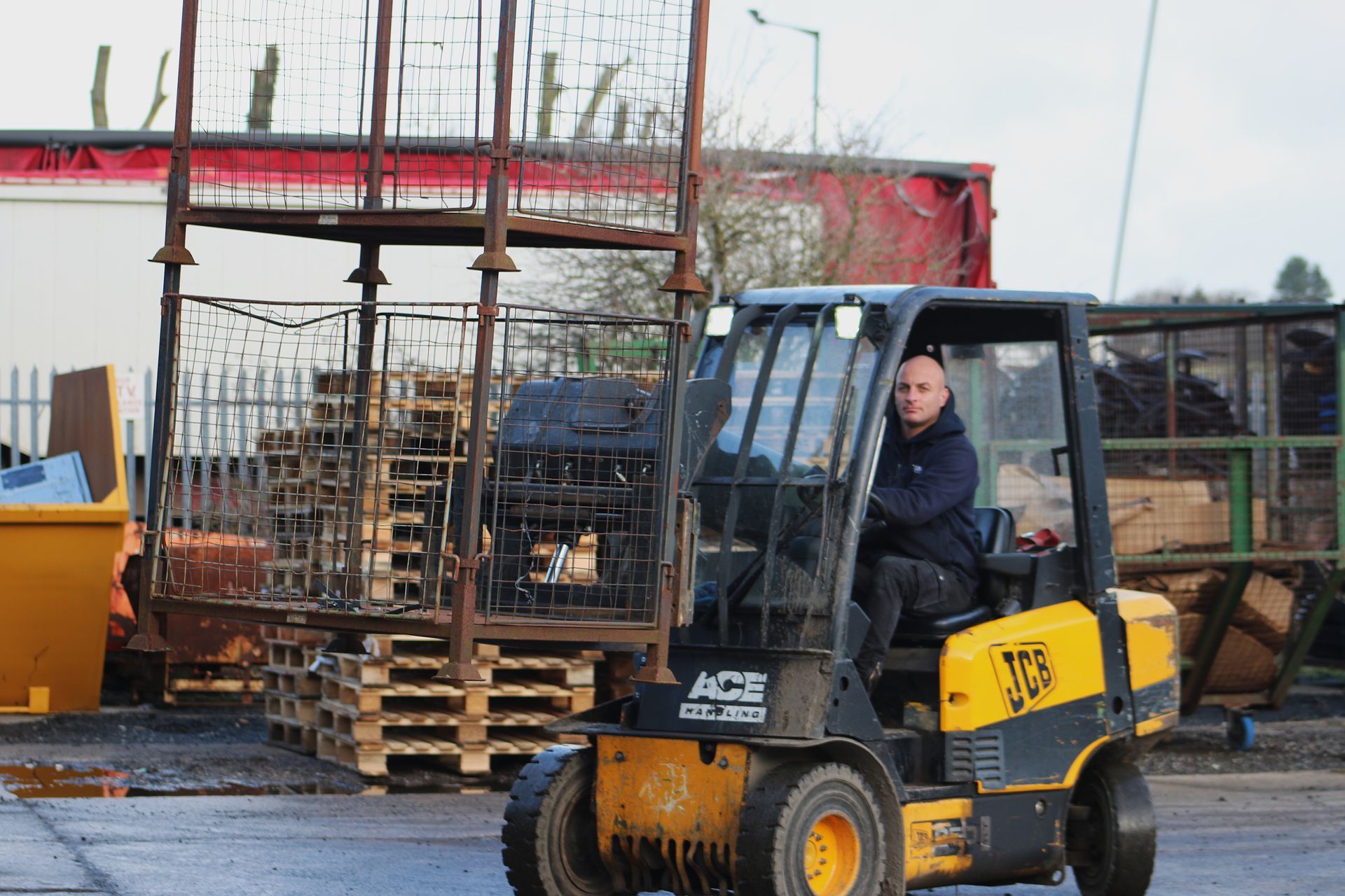 Man driving a yellow JCB forklift, lifting a metal cage in an industrial yard.