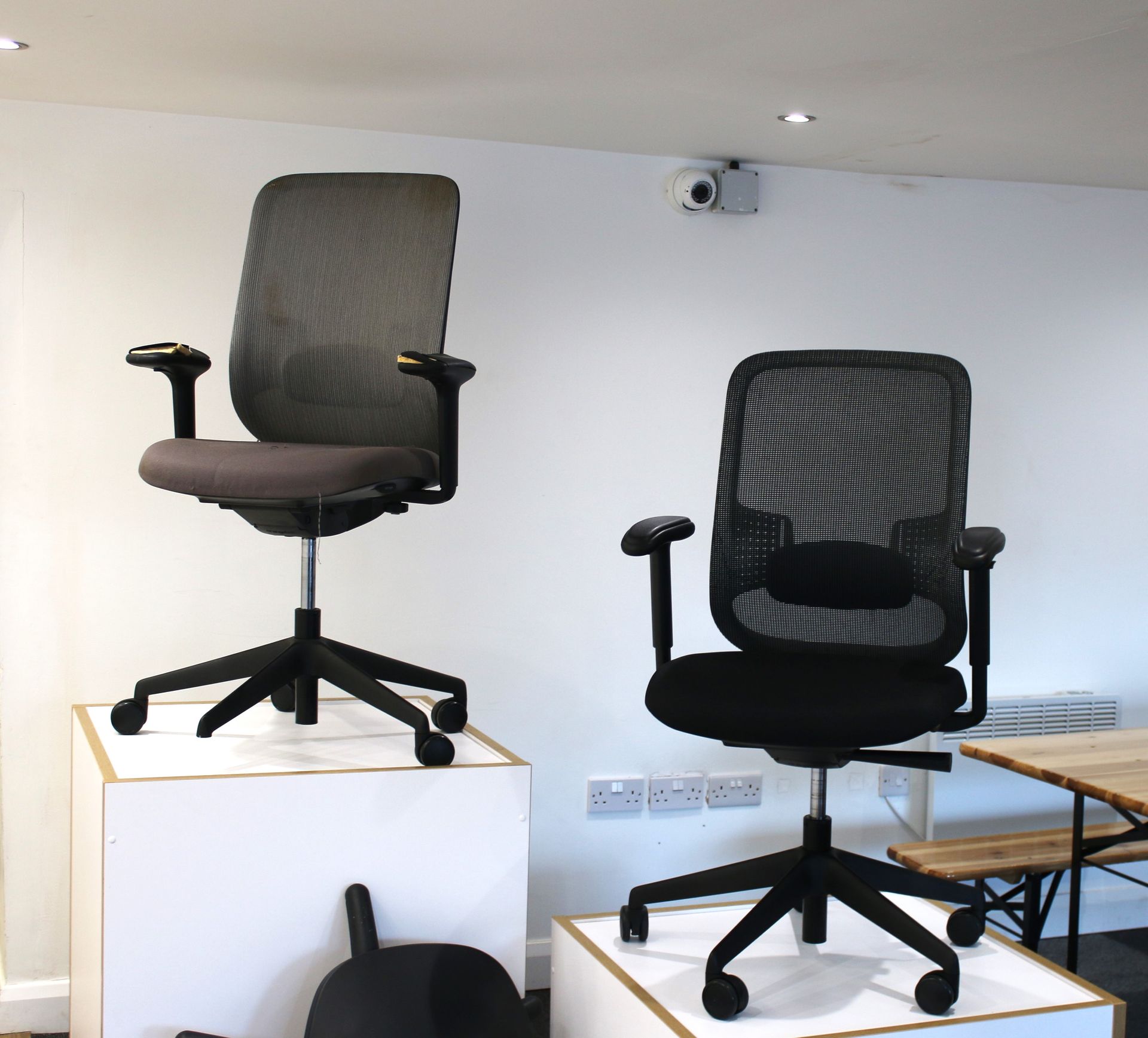 Two mesh office chairs on white platforms, against a white wall.