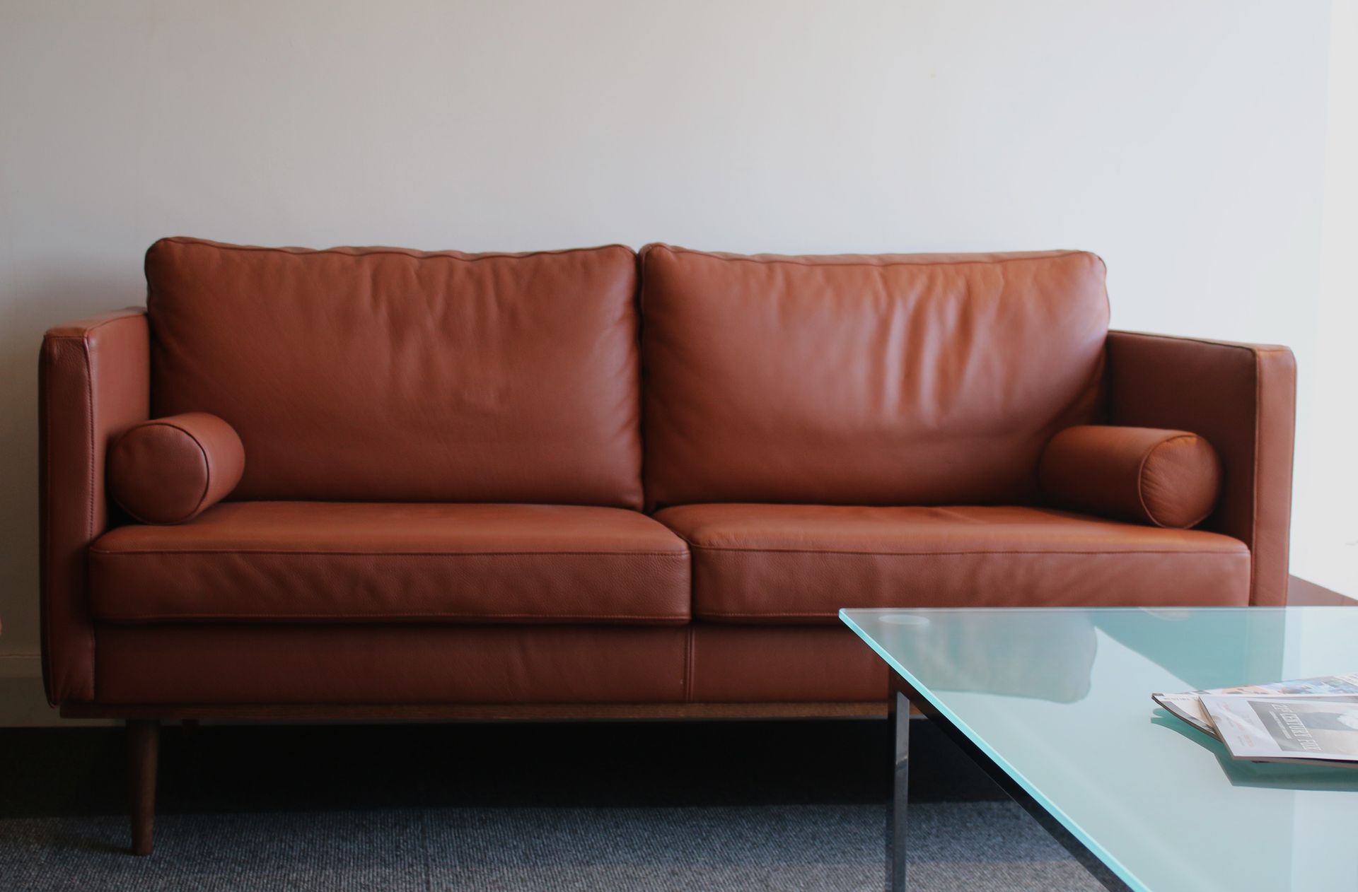 Brown leather sofa with two cushions and a glass coffee table in front of a white wall.