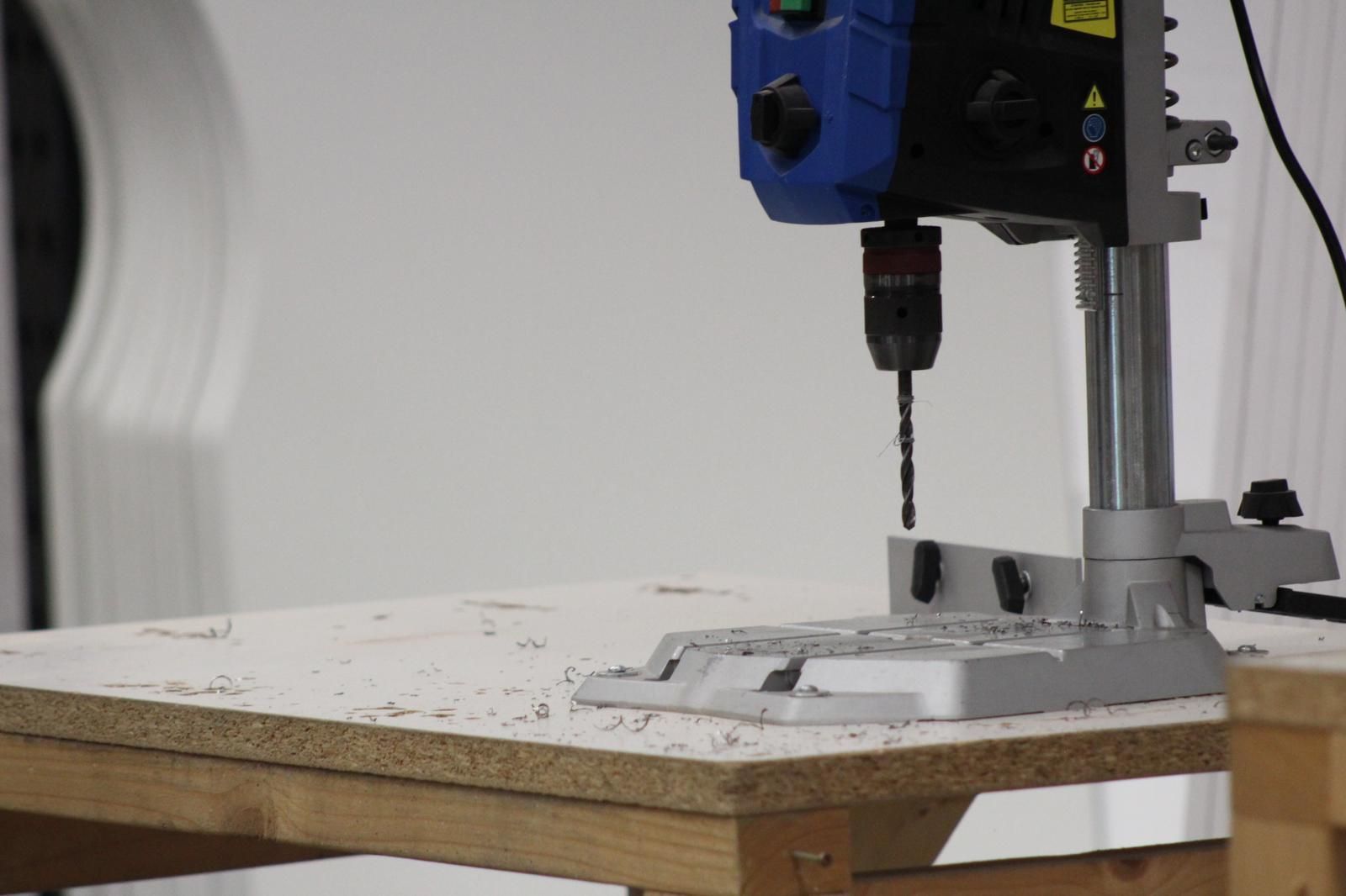 A drill press, blue and black, drilling into a wood surface; wood shavings visible.